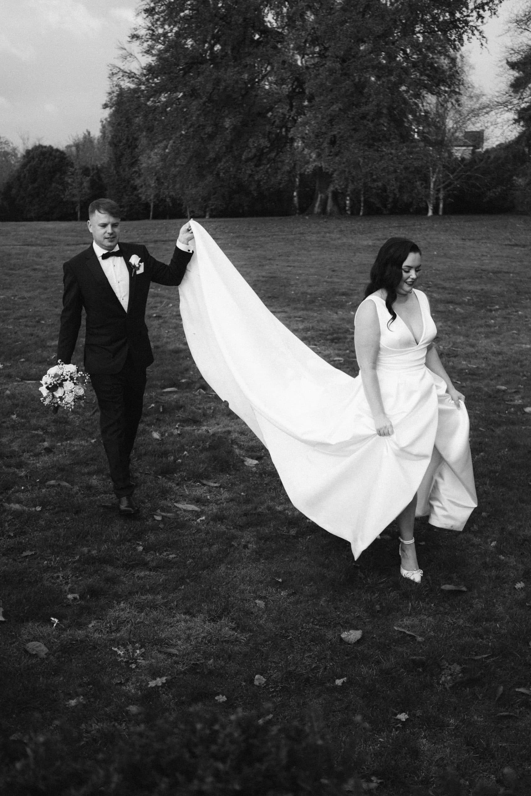 Groom carries his wife's dress as they walking in the gardens at Swynford Manor